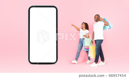 Excited black couple with shopping bags walking near giant smartphone with empty screen, pink background, mockup 98957454