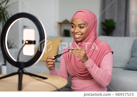 Beauty blogger. Happy african american lady in hijab reviewing new mascara, looking at smartphone set on ring lamp Beauty blogger. Happy african american lady in hijab reviewing new mascara, looking at smartphone set on ring lamp 98957523