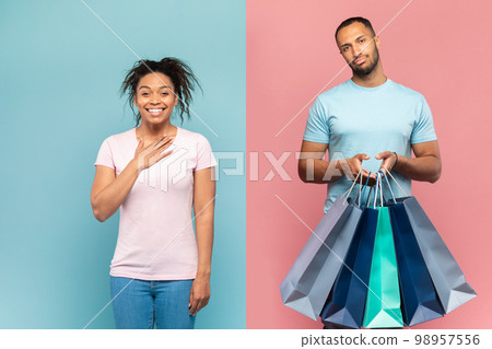 Displeased black guy standing with shopper bags, shopping with his overjoyed lady, standing on pink and blue background 98957556