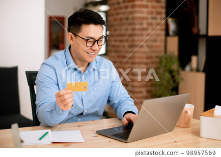Fast online shopping. Happy asian businessman holding debit credit card and using laptop, making financial transaction Fast online shopping. Happy asian businessman holding debit credit card and using laptop, making financial transaction 98957569