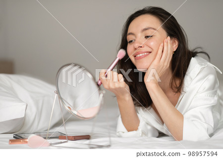 Cheerful caucasian millennial woman applies powder with brush on face, looks in mirror, enjoy good morning 98957594