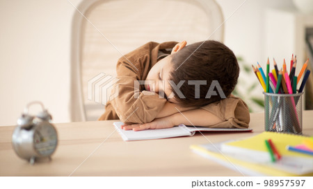 Bored unhappy tired caucasian little child sleeping at table, suffering from homework in room Bored unhappy tired caucasian little child sleeping at table, suffering from homework in room 98957597