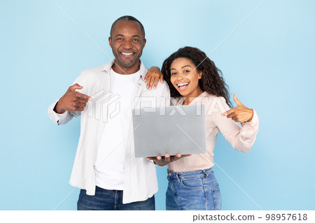 Happy african american couple holding laptop and pointing fingers at gadget, standing over blue studio background Happy african american couple holding laptop and pointing fingers at gadget, standing over blue studio background 98957618