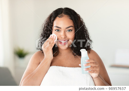 Beautiful black body positive lady cleaning her face from makeup with cotton pads and micellar water, home interior 98957637