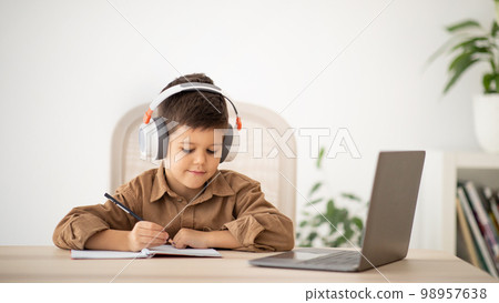 Smiling caucasian little kid in headphones sits at table, makes notes, watches lesson on computer in room 98957638