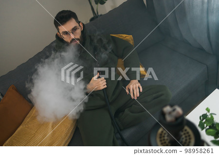 Bearded millennial or gen z man smoking hookah while relaxing on sofa at home - chill time and resting concept 98958261