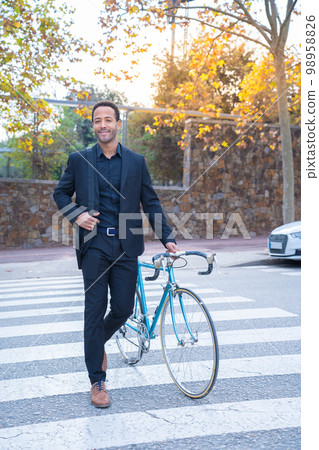 Stylish African American businessman in suit crossing the street with the bicycle. Stylish African American businessman in suit crossing the street with the bicycle. 98958826