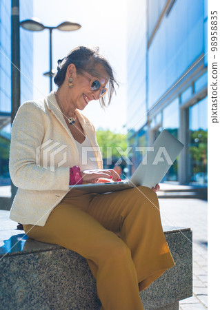 Happy attractive business woman middle-aged working smiling with laptop in front of a modern building. Vertical photo. 98958835