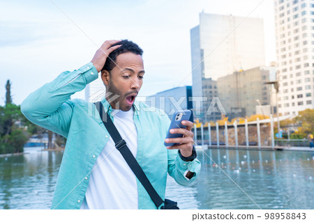 Young black man in the street looking at the phone with a surprised expression. 98958843