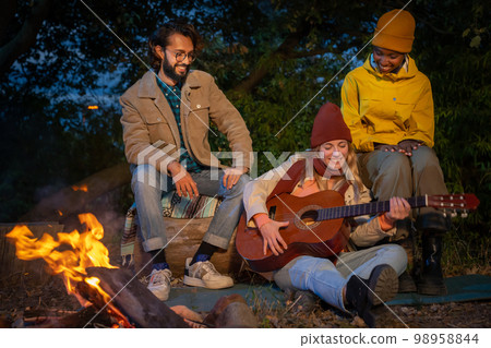 Three happy diverse friends having fun playing music and enjoying bonfire camping in nature at night 98958844
