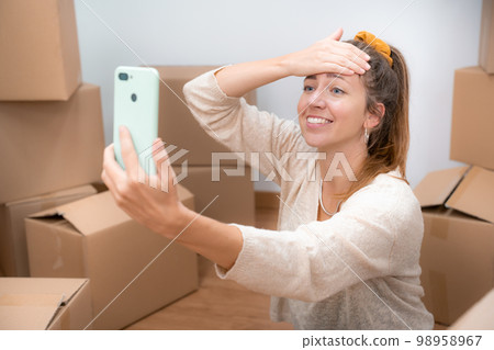 Beautiful girl with cardboard boxes unpacking in new home taking a selfie or video call. 98958967