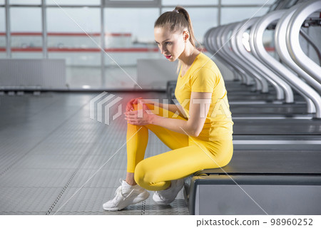Young woman in sportswear having pain in her knee while training in gym, Girl sitting on a floor touching her knee in pain 98960252