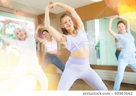 Group of young mature emotional female dancers raising both hands and having energetic training in dance hall Group of young mature emotional female dancers raising both hands and having energetic training in dance hall 98963662