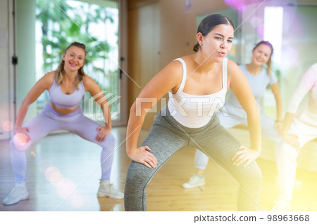 Cheerful young mature female dancers in sportswear dancing with legs wide apart during exercising with coach in gym 98963668