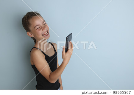 Little emotional teenage girl in pink shirt 11, 12 years old on an isolated blue background with a tablet in her hands.Children's studio portrait. Place the text to copy the place for the inscription 98965567