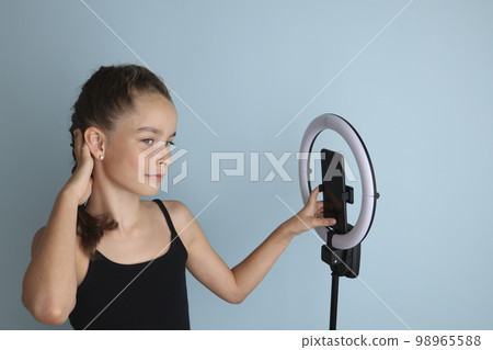 Little emotional teenage girl in a black t-shirt on an isolated blue background. Studio portrait of a young smiling teenage girl, vlogger, with a ring flash lamp and a smartphone. Girl takes a selfie. Little emotional teenage girl in a black t-shirt on an isolated blue background. Studio portrait of a young smiling teenage girl, vlogger, with a ring flash lamp and a smartphone. Girl takes a selfie. 98965588