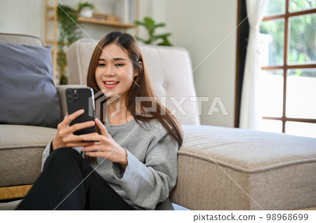 Relaxed Asian female uses her smartphone to chat with her friends in living room. Relaxed Asian female uses her smartphone to chat with her friends in living room. 98968699