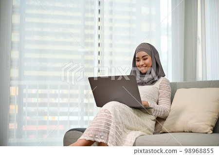 Elegant Asian Muslim woman using laptop to manage her tasks while relaxing on sofa 98968855