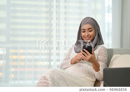 Attractive Asian Muslim pregnant woman resting, using her mobile phone on the sofa. 98968856