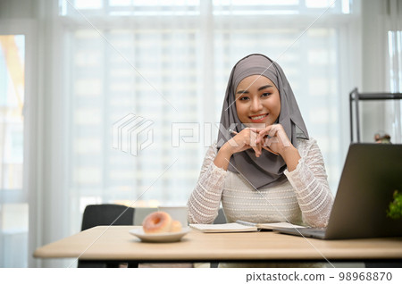 Gorgeous Asian Muslim businesswoman sitting at her office desk, smiling and looking at the camera. 98968870