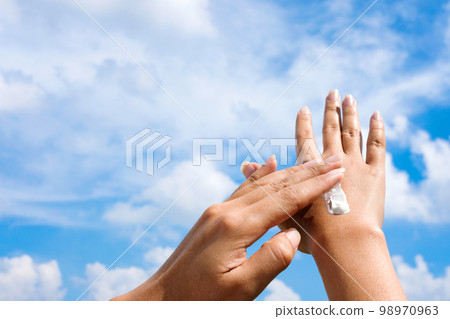 A woman is applying sunscreen and skin care to protect her skin from UV rays. She is applying sunscreen on her hand and arm. The sun symbol is a very sunny background. Health and skin care concept A woman is applying sunscreen and skin care to protect her skin from UV rays. She is applying sunscreen on her hand and arm. The sun symbol is a very sunny background. Health and skin care concept 98970963