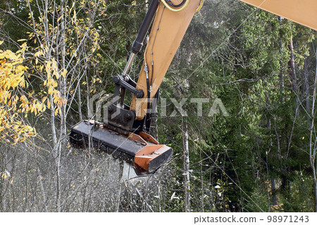 Mulching vegetation on side of road in wooded area with an excavator with mulching head. Mulching vegetation on side of road in wooded area with an excavator with mulching head. 98971243
