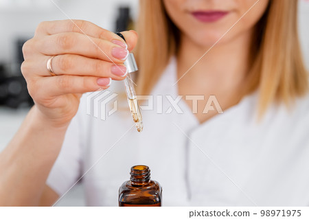 Woman drops of a crystal clear cosmetic from pipette in glass jar. 98971975