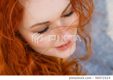 Close-up portrait young caucasian woman with red curly hair in red coat in wind 98972824