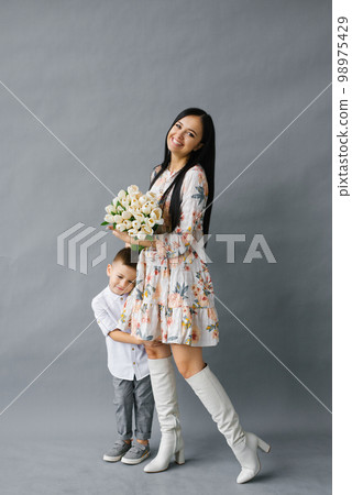 Photo portrait of a brunette girl holding a bouquet of white tulips in her hands, standing next to her four-year-old child, isolated against a gray background. Mother's Day 98975429