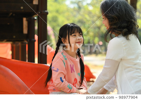 Happy child girl on slide, having fun on outdoor playground with grandma. Childhood and equipment of entertainment park Happy child girl on slide, having fun on outdoor playground with grandma. Childhood and equipment of entertainment park 98977904