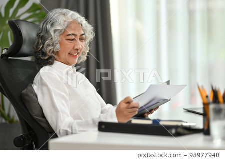 Pleasant middle age businesswoman business women reading analysis graph in paper document while sitting on office chair Pleasant middle age businesswoman business women reading analysis graph in paper document while sitting on office chair 98977940