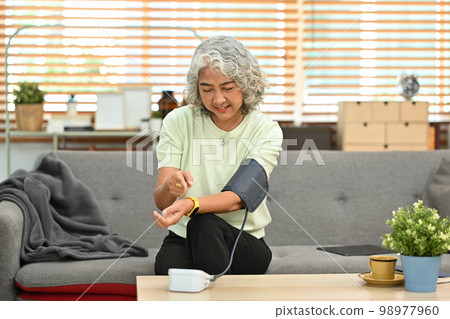 Senior woman checking blood pressure, sitting at a couch and using digital tonometer. Health and Medical concept Senior woman checking blood pressure, sitting at a couch and using digital tonometer. Health and Medical concept 98977960