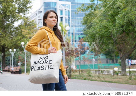 A brunette woman in an orange hoodie walks with a fabric bag that says No more plastic in the city 98981374