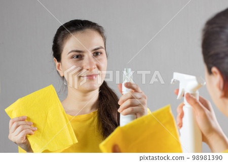 A beautiful woman in a yellow bright T-shirt smiles and cleaning mirror with a water squeegee with a scraper. Means for cleaning windows and glass. Cleaning service. Maid cleaning at home. 98981390