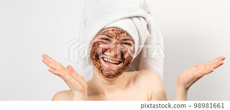 A young woman with a coffee mask on her face and a bath towel on her head smiles and looks into the camera on a white background, a place for text. 98981661