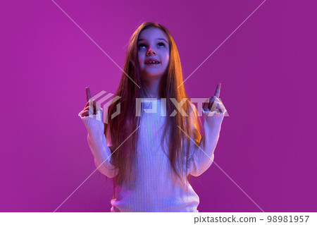 Dreaming. Portrait of little girl, child in casual clothes posing, raising two fingers up over purple studio background in neon light. Concept of emotions, childhood Dreaming. Portrait of little girl, child in casual clothes posing, raising two fingers up over purple studio background in neon light. Concept of emotions, childhood 98981957