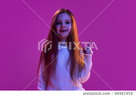 Ideas. Portrait of little girl, child in casual clothes posing, raising finger up over purple studio background in neon light. Concept of emotions, childhood 98981958