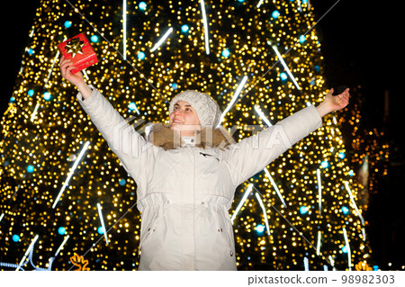 A young woman in a white jacket and hat holds the gift standing near the Christmas tree, raising her hands up A young woman in a white jacket and hat holds the gift standing near the Christmas tree, raising her hands up 98982303