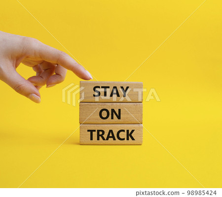 Stay on track symbol. Wooden blocks with words 'Stay on track'. Beautiful yellow background. Businessman hand. Business and 'Stay on track' concept. Copy space Stay on track symbol. Wooden blocks with words 'Stay on track'. Beautiful yellow background. Businessman hand. Business and 'Stay on track' concept. Copy space 98985424