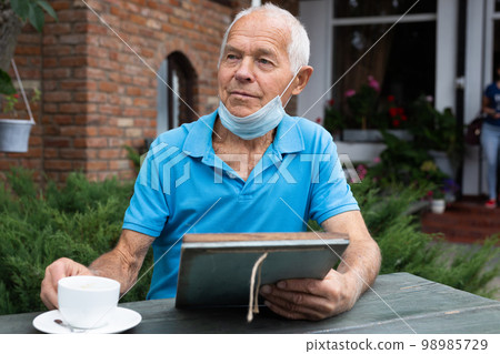 Senior man in protective mask chooses dish from the menu while sitting at the table of street cafe Senior man in protective mask chooses dish from the menu while sitting at the table of street cafe 98985729