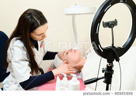 A beautician performs a face massage by rubbing cream into the skin, an LED lamp is in the foreground. 98985898