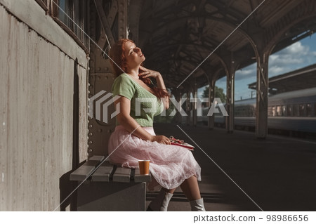 redhead young woman in green bun and pink skirt dreamily sits with glass of coffee at train station 98986656