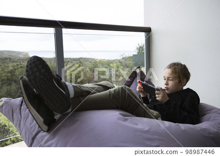 Teenage boy enjoying his free time and playing game on handheld console. Child lying on beanbag bed on the balcony with nature view Teenage boy enjoying his free time and playing game on handheld console. Child lying on beanbag bed on the balcony with nature view 98987446