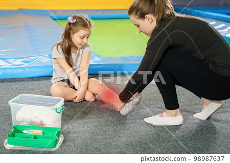 Little girl having pain in ankle and getting help after jumping on trampoline, red spot showing ache 98987637