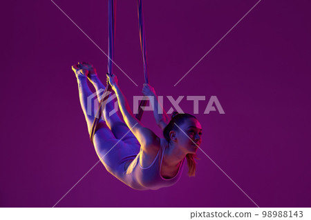 Relaxing activity. Young flexible girl doing aerial yoga, training over purple studio background in neon light. Concept of fitness and healthy lifestyle 98988143