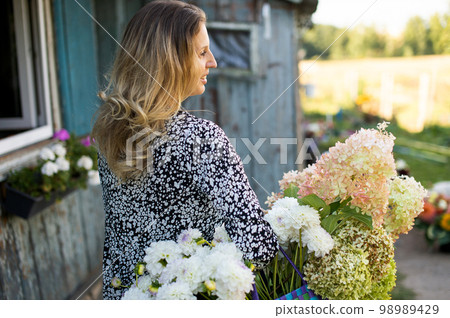 A girl in a dress with a basket of dahlia and hydrangea flowers passes near a house in the village A girl in a dress with a basket of dahlia and hydrangea flowers passes near a house in the village 98989429