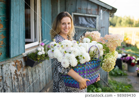 A girl in a dress with a basket of white dahlia flowers stands near a house in the village A girl in a dress with a basket of white dahlia flowers stands near a house in the village 98989430
