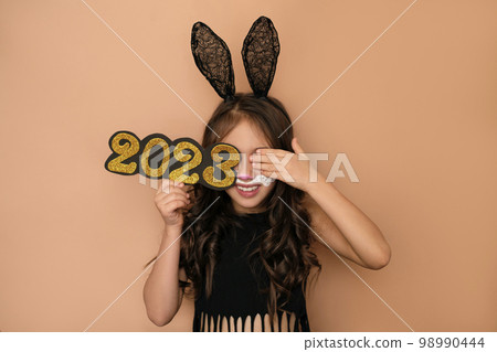A beautiful girl dressed up as a rabbit holds the figure of the new year 2023 and closes her eye with her hand laughing merrily. Lack of confidence in the future and fear 98990444