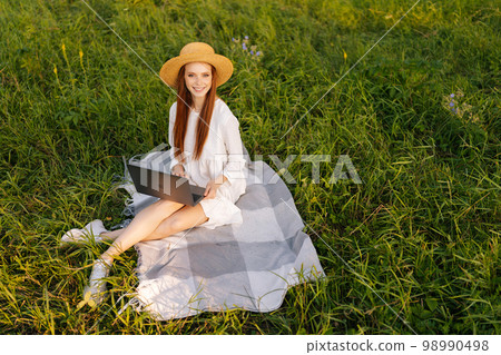 Top view of gorgeous positive young woman wearing straw hat and white dress sitting posing on plaid in with laptop computer looking at camera, on beautiful field of green grass on summer day. Top view of gorgeous positive young woman wearing straw hat and white dress sitting posing on plaid in with laptop computer looking at camera, on beautiful field of green grass on summer day. 98990498