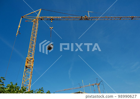 A tall yellow construction crane with a dangling black box against a blue sky in Germany. A tall yellow construction crane with a dangling black box against a blue sky in Germany. 98990627